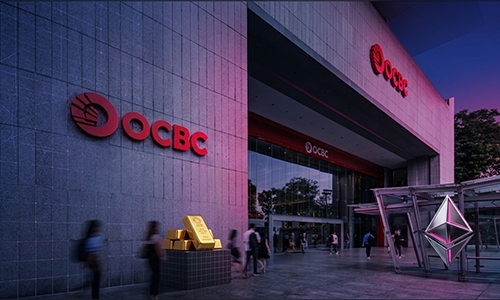 OCBC bank exterior at dusk with red logo, glass entrance, and a display of gold bars outside.