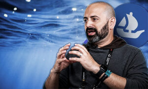 Bearded man in a dark shirt gesturing while speaking at a conference, wearing a lanyard and smartwatch, against a blue backdrop with a sail logo.